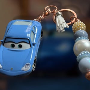 May include: A light blue car-shaped keychain with a beaded accent. The keychain features a small, light blue car with black detailing and a cartoon face. The beaded accent includes white, blue, and gold beads, and a white tassel.