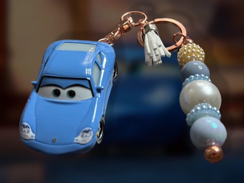 May include: A light blue car-shaped keychain with a beaded accent. The keychain features a small, light blue car with black detailing and a cartoon face. The beaded accent includes white, blue, and gold beads, and a white tassel.
