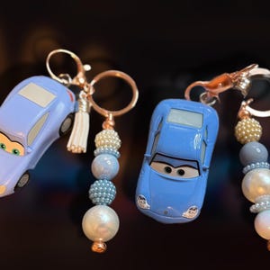 May include: Two car-shaped keychains. One is light purple, the other is blue. Each keychain has a gold-colored ring and a beaded charm with pearls and blue beads. One has a white tassel.