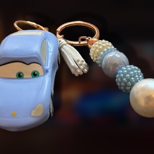 May include: A light blue car-shaped keychain with a gold-colored keyring and a white tassel. The keychain features a beaded accent in shades of blue, white, and pearl. The car has green eyes and a smiling face.