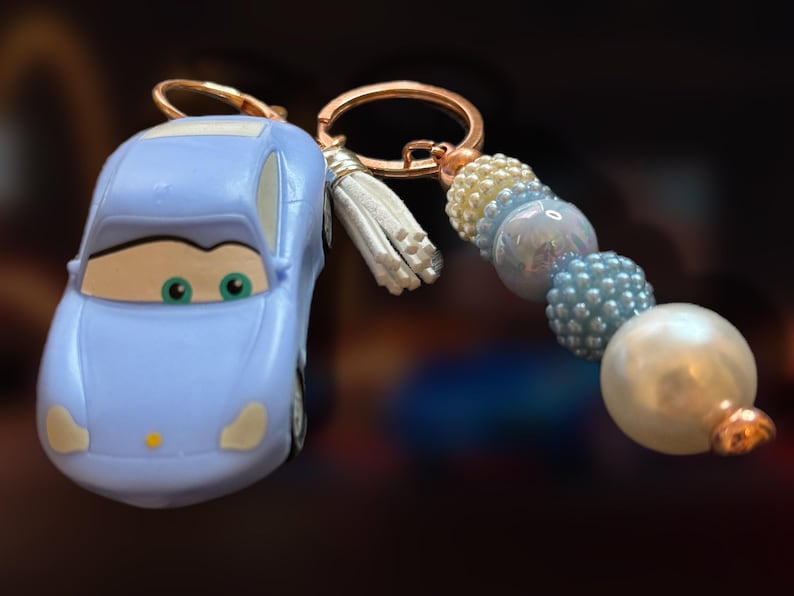 May include: A light blue car-shaped keychain with a gold-colored keyring and a white tassel. The keychain features a beaded accent in shades of blue, white, and pearl. The car has green eyes and a smiling face.