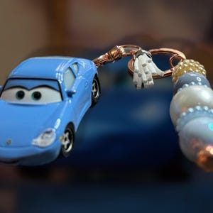 May include: A light blue toy car keychain featuring a cartoon car design. The keychain includes a decorative beaded charm with white, blue, and gold beads, and a small white tassel. The car has black wheels and a smiling face.