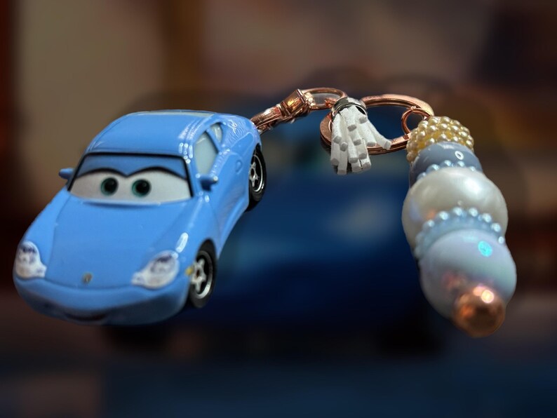 May include: A light blue toy car keychain featuring a cartoon car design. The keychain includes a decorative beaded charm with white, blue, and gold beads, and a small white tassel. The car has black wheels and a smiling face.