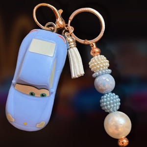 May include: A light blue car-shaped keychain with a gold-colored clasp and a white tassel. The keychain features a beaded accent in shades of white, blue, and pearl, with a gold-colored bead at the bottom.