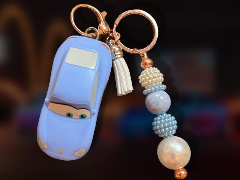 May include: A light blue car-shaped keychain with a gold-colored clasp and a white tassel. The keychain features a beaded accent in shades of white, blue, and pearl, with a gold-colored bead at the bottom.