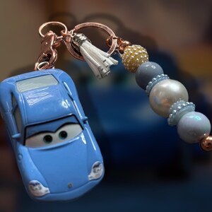 May include: A light blue car-shaped keychain with a cartoon face, attached to a rose gold keyring. The keychain features a white tassel and a beaded strand with gold, gray, and pearl-like beads. A fun accessory.