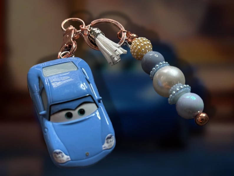 May include: A light blue car-shaped keychain with a cartoon face, attached to a rose gold keyring. The keychain features a white tassel and a beaded strand with gold, gray, and pearl-like beads. A fun accessory.