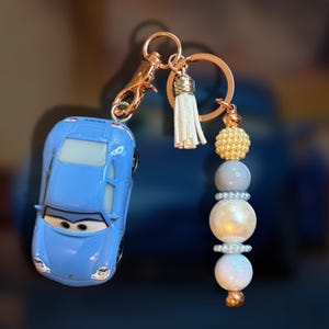 May include: A blue toy car keychain with a gold-colored clasp and ring. The keychain features a white tassel and a beaded charm with gold, gray, and white beads. The car has a cartoon face.