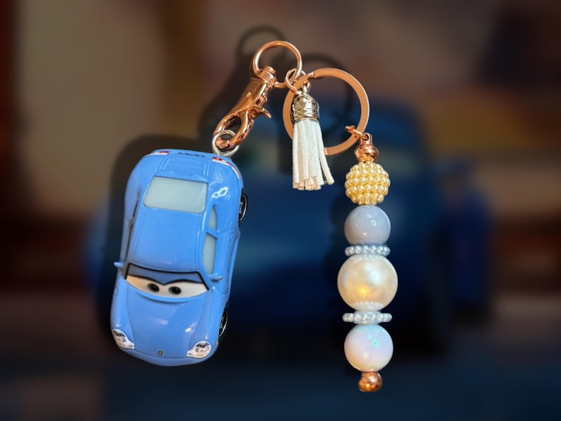 May include: A blue toy car keychain with a gold-colored clasp and ring. The keychain features a white tassel and a beaded charm with gold, gray, and white beads. The car has a cartoon face.