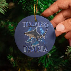 May include: A round, navy blue Christmas ornament with a shark wearing blue sneakers and the text "TRALALERO TRALALA". The ornament is hanging on a green Christmas tree with red ornaments and a red and white striped ribbon.