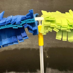 May include: A two-sided cleaning duster with a long handle. One side is blue, the other is green. The duster has a yellow and white handle. The cleaning heads are made of soft, fringed material.
