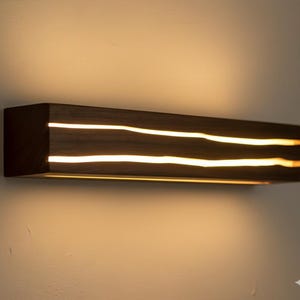 Op de afbeelding: Een rechthoekige, donkerbruine houten wandlamp met twee horizontale, gloeiende, golvende lijnen van warm wit licht. De armatuur is gemonteerd op een lichtbeige muur, waardoor een contrast ontstaat.