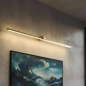 Può includere: Un moderno apparecchio di illuminazione a parete rettangolare con una calda luce bianca. L'apparecchio ha una finitura oro spazzolato ed è montato su una parete grigia strutturata sopra un dipinto incorniciato con toni blu e bianchi.