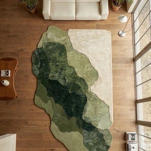 Könnte beinhalten: Draufsicht auf ein modernes Wohnzimmer mit einem großen, abstrakten Teppich in Grün- und Beigetönen. Das Design des Teppichs ähnelt einer Landschaft. Ein cremefarbenes Sofa, Pflanzen und ein Holzboden ergänzen die Szene.