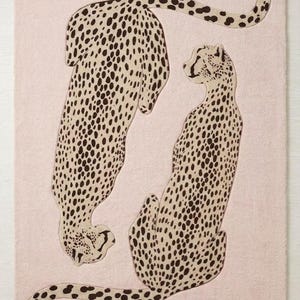 May include: A rectangular rug with a pale pink background features two cheetahs. The cheetahs are depicted in a beige colour with dark brown spots. One cheetah is lying down, and the other is sitting upright.