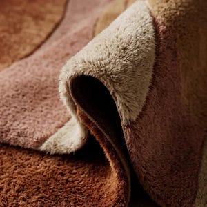 May include: Close-up of a plush, abstract rug with a rolled edge. The rug features a pattern of overlapping shapes in shades of brown, tan, and pink. The texture appears soft and inviting, suitable for a living room or bedroom.