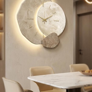 Può includere: Un orologio da parete moderno con un design unico. L'orologio presenta un quadrante circolare con lancette dorate e un aspetto strutturato, simile alla pietra. L'orologio è circondato da una forma geometrica irregolare ed è illuminato da una luce calda e soffusa. L'orologio è montato su una parete di colore neutro.