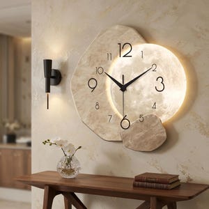 Puede incluir: Un reloj de pared moderno con un diseño único. El reloj presenta una forma de piedra clara e irregular con un círculo brillante en forma de luna en el centro. Las manecillas y los números negros del reloj son visibles. Una lámpara de pared negra está a la izquierda.