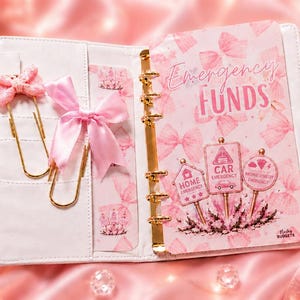 May include: A white six-ring binder featuring a pink bow pattern and a dashboard labeled Emergency Funds with icons for home, car, and miscellaneous emergencies. Includes two gold jumbo paperclips with pink bows and a matching pen with a bow topper.