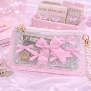May include: A clear, pink-trimmed pouch with pink bows and a gold bow clasp. The pouch contains money and coins, and is attached to a pearl wristlet. The background includes a pink notepad, pens, and a calculator.