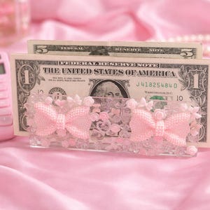 May include: A pink calculator and a decorative money holder with two pink bows and floral accents hold US dollar bills. The items are set against a pink satin background, creating a soft, feminine aesthetic.