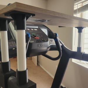 Könnte beinhalten: Ein Holzschreibtischaufsatz für ein Laufband. Der Schreibtisch wird von schwarzen und weißen Pfosten getragen und ist an der Konsole des Laufbands befestigt. Das Laufband hat ein digitales Display und blaue Griffe. Der Schreibtisch ist aus hellbraunem Holz.