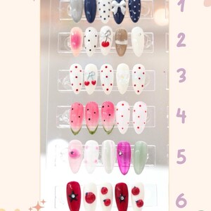 May include: A display of artificial nails in various designs. The nails feature colors like gray, navy, white, pink, and green, with patterns including polka dots, bows, cherries, and watermelon slices. Some nails have embellishments like rhinestones.