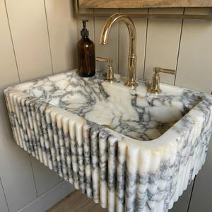 May include: A marble sink with a fluted design and a gold tap. The sink features a white and grey veined pattern. A brown glass bottle with a black pump sits on the sink's surface.