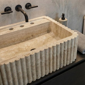 May include: A beige stone rectangular sink with a fluted design. The sink has a dark faucet and handles, with a matching soap dispenser and a gray towel nearby. The sink is set against a gray wall.
