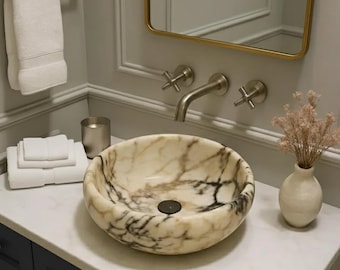 May include: A round marble sink with brown veining is set on a white countertop. A brushed nickel tap and cross-handle taps are mounted on the wall. White towels hang on a rail, and a small vase holds dried flowers.