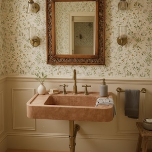 May include: A bathroom interior with a peach-toned sink and brass taps, set against floral wallpaper and a decorative wooden mirror. A soap dispenser, small vase, and towel rail are visible. Wall sconces provide lighting.
