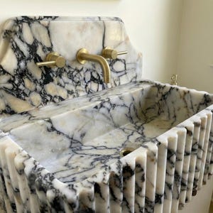 May include: A marble sink with a white and black veined pattern. The sink has a rectangular basin with a fluted front and a backsplash with a curved top. Gold-colored faucet and handles are mounted on the backsplash.