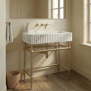 以下が含まれることがあります： 垂直の溝が入った白い大理石の長方形の洗面台。金色の金属フレームの上に設置されています。洗面台には内蔵の蛇口があります。金色のタオル掛けと籐のバスケットも写っています。