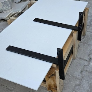 May include: A white rectangular shelf with two black metal brackets attached. The shelf is supported by a wooden structure. The brackets are mounted on the underside of the shelf and are designed to be mounted on a wall.