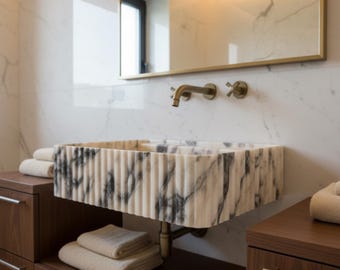 May include: A modern bathroom sink made of white marble with black veining. The sink has a rectangular shape with vertical ribbed detailing. A gold-toned faucet and mirror frame complement the sink. The sink is mounted above a dark wood cabinet with towels.