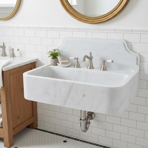 May include: A white marble bathroom sink with a curved front and a backsplash. The sink has a brushed nickel tap and white porcelain handles. A small potted plant and soap dish sit on the sink's surface. The sink is mounted on a white tiled wall.