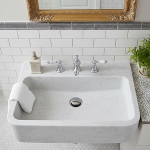 May include: A white marble bathroom sink with a chrome faucet and white porcelain handles. A white hand towel rests on the sink's edge. A marble soap dispenser and a potted plant are also visible. The sink is set against a white tiled wall.
