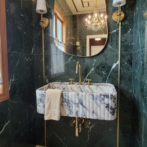 May include: A luxurious bathroom with a marble sink and gold fixtures, a large oval mirror, and a crystal chandelier. The walls are covered in dark green marble. Two gold sconces flank the mirror, and a beige towel hangs on the sink.