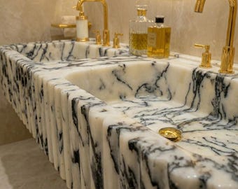 May include: A double sink made of white marble with black veining. The sink has a fluted edge and gold-colored faucets and soap dispensers. Bottles of liquid are on the countertop.