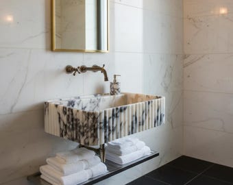 May include: A modern bathroom with a marble sink featuring vertical ribbed detailing and a black and white pattern. A stainless steel shelf holds folded white towels. The walls are white marble, and the floor is black tile.