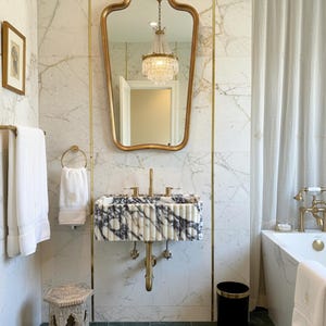 May include: Luxurious bathroom featuring marble walls and a black and white marble sink. A gold-framed mirror reflects a chandelier. White towels and a small stool enhance the space. A black bin with a gold rim is also visible.