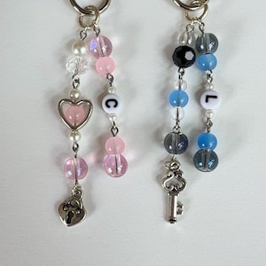 May include: Two beaded keychains with silver-tone hardware. One features pink and white beads, a heart charm, and a lock charm. The other has blue, black, and gray beads, a key charm, and the letter "L".