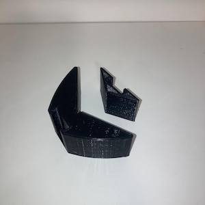 May include: Two black 3D-printed objects. One is a curved, open-topped container with a pointed side. The other is a smaller, angular piece with a slot. Both objects are on a white surface.