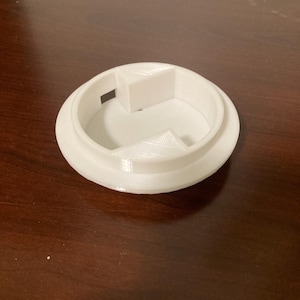 Puede incluir: Un objeto blanco impreso en 3D con una base circular y un borde redondeado elevado. En el interior, hay un área central empotrada con un pequeño bloque rectangular y una estructura de soporte triangular. El objeto es probablemente una tapa.