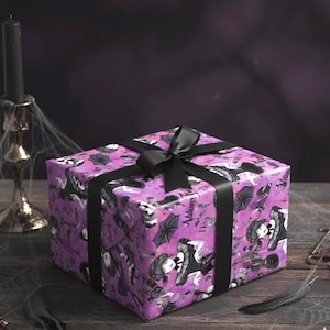 Puede incluir: Una caja de regalo envuelta con un lazo negro. El papel de regalo es morado con un patrón en blanco y negro con ilustraciones de una mujer, paraguas y el texto "Addams". Una cinta negra está atada alrededor de la caja.