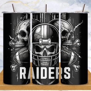 Könnte beinhalten: Schwarz-weißer Becher mit einem Totenkopf, der einen Footballhelm trägt, flankiert von zwei Totenköpfen und Fußbällen. Das Wort "RAIDERS" ist unten in fetten weißen Buchstaben aufgedruckt. Enthält einen Strohhalm und einen schwarzen Deckel.