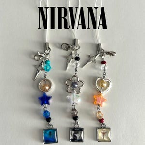 May include: Three beaded phone charms with silver-tone hardware. Each charm features a different design: a star, a flower, and a heart. The charms include various beads in blue, clear, and orange, and small square pendants with images. The word "NIRVANA" is at the top.