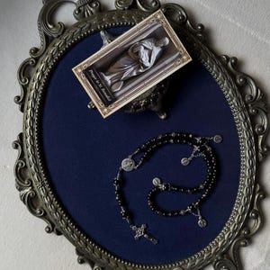 May include: An ornate, oval-shaped, antique-style mirror frame with a dark blue interior. A black rosary and a small card with the text "Prayer to St. Benedict" are displayed inside the frame. The rosary features a cross and silver-toned accents.