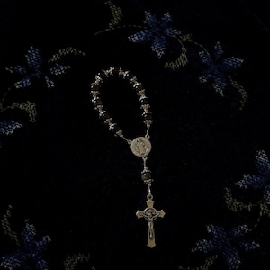 May include: A silver rosary with black beads and a crucifix. The rosary is set against a dark background with blue floral embroidery. The crucifix features a detailed depiction of Jesus Christ.