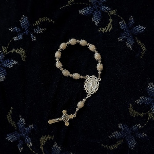 May include: A rosary with pale pink beads and silver-toned metal accents. The rosary features a crucifix and a centerpiece with a religious figure. The beads are strung together with delicate metal links.